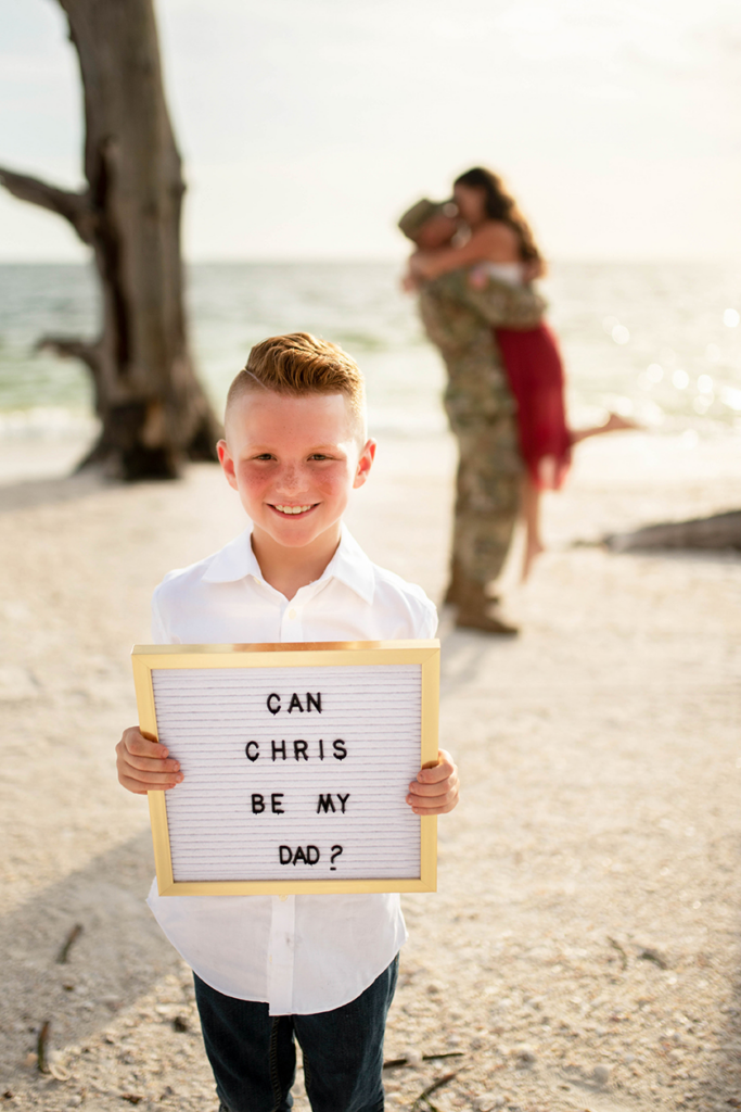 A boy stands on the beach as his parents kiss behind him, holding a sign that says "can Chris be my dad?". The engaged couple in the background embraces and shares a kiss.