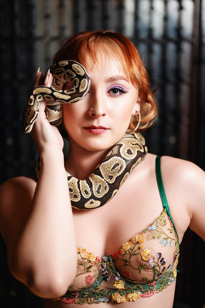 a woman in garden-themed lingerie bra has a ball python wrapped around her neck, over her head, and covering one eye.