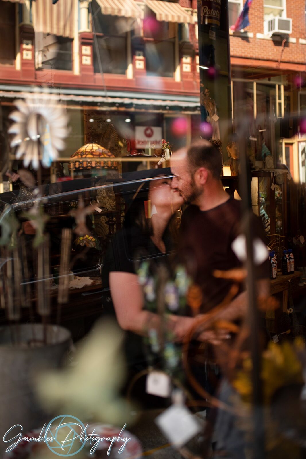 with the shining lights of Eureka Springs reflecting on the storefront's window, a couple stands inside and shares a kiss. Photo by Gambles Photography