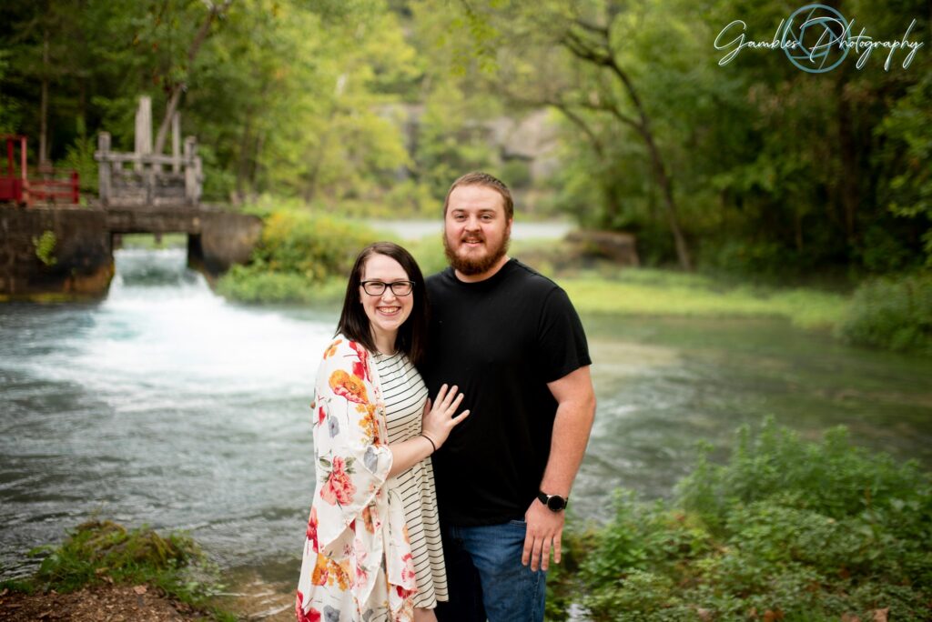 A hand on her fiancee's chest, a couple smiles near idyllic blue water and a towering red barn after their proposal at Alley Springs and Mill. Photo by Gambles Photography