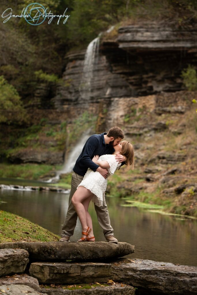 Kissing his fiancee as she leans backwards, a couple stands at Dogwood Canyon Nature Park after their engagement, with mossy rocks, still water, and a waterfall behind them. Photo by 