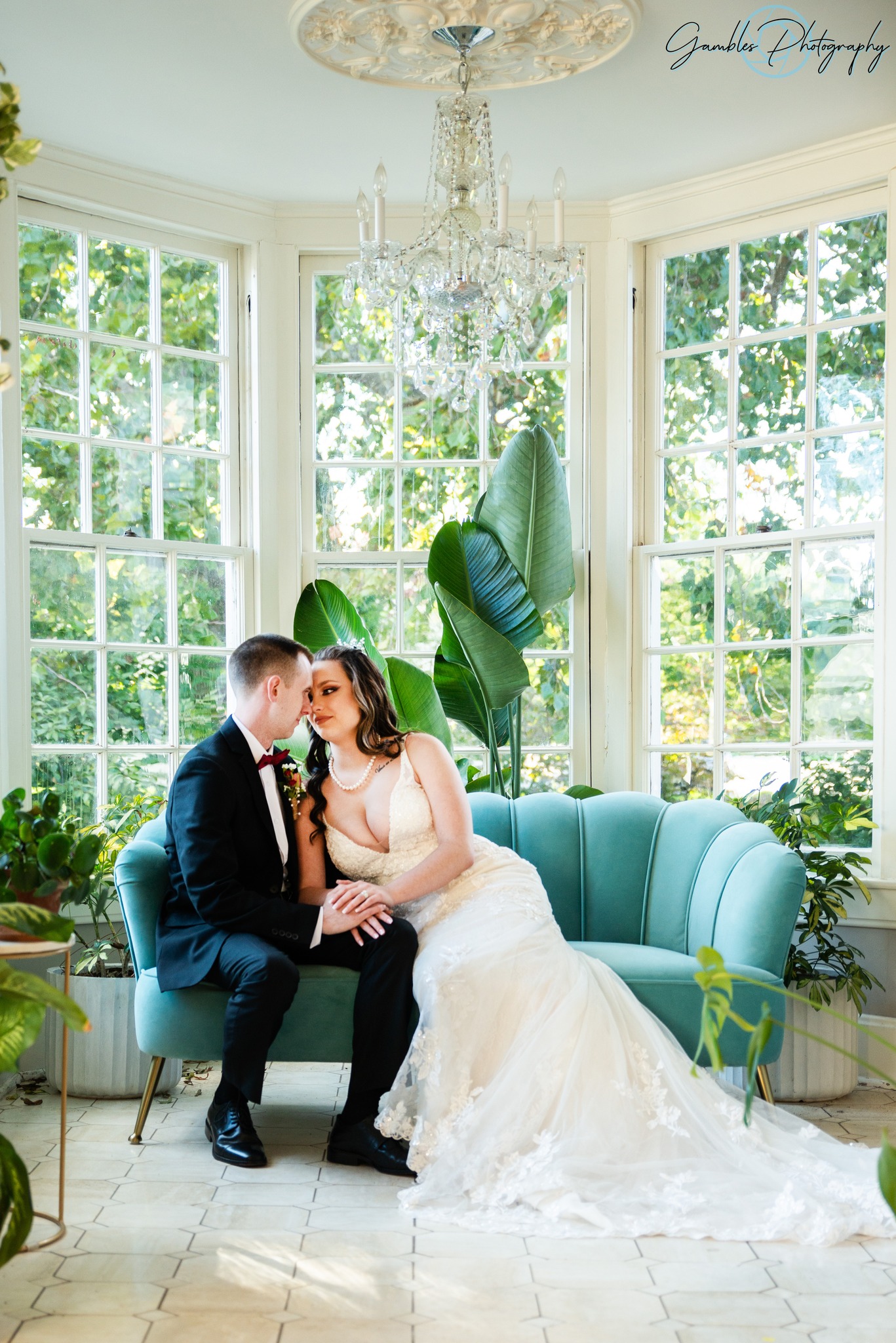 In front of large bay windows and sitting on a velvet, turquoise couch, a bride leans in to kiss her husband on their wedding day at Haseltine Estate in Springfield, Missouri