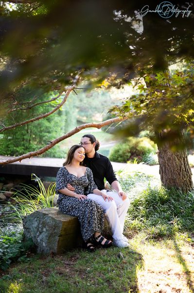 A couple sits on a rock after their proposal at Mizumoto Japanese Stroll Garden, photographed by Gambles Photography. The dappled sunlight shines through the trees, as the man kisses the top of his soon-to-be wife's head.