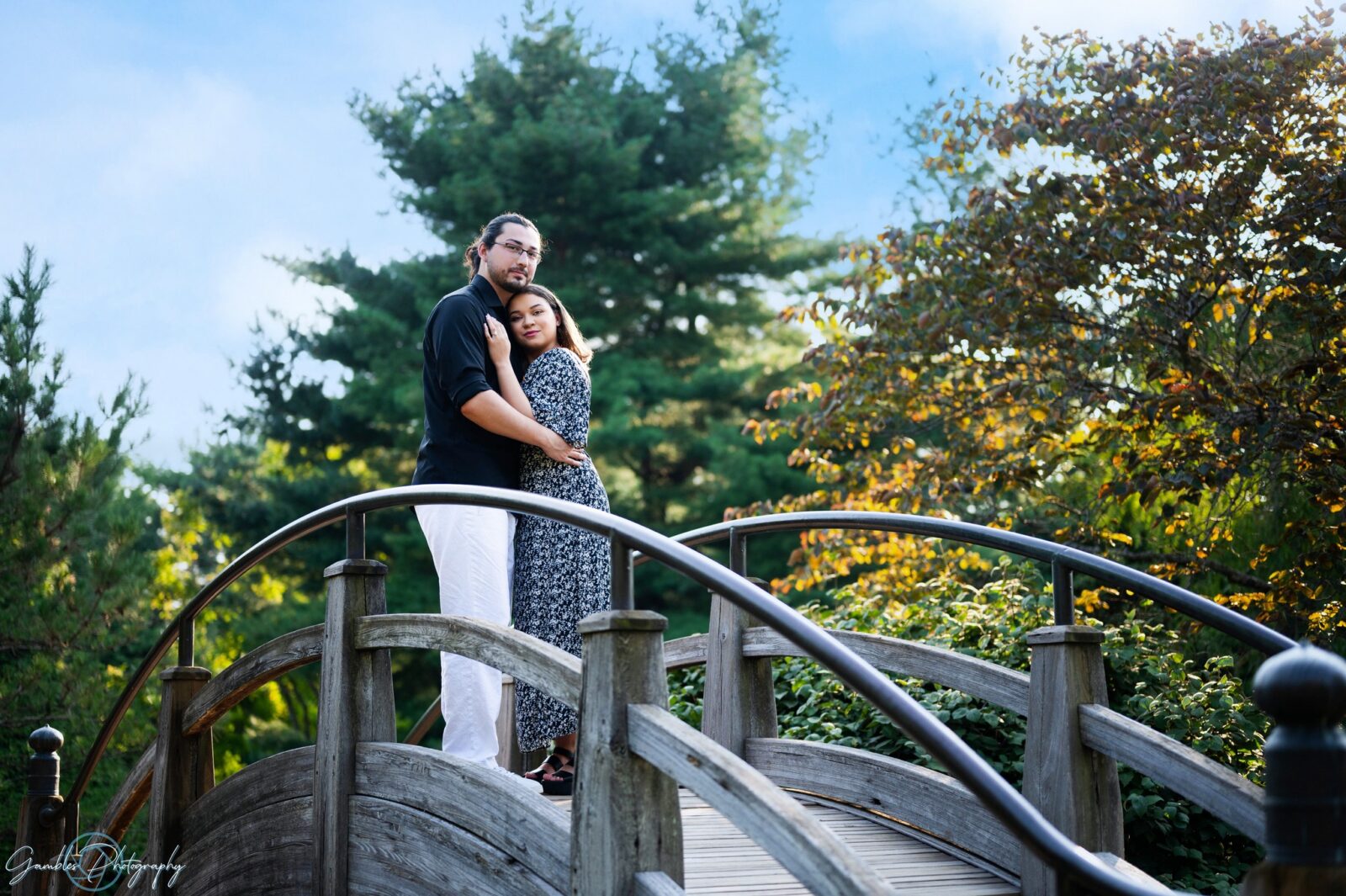 A couple embraces atop a small, arched wooden bridge at Mizumoto Japanese Stroll Garden in Springfield, MO. By Gambles Photography.