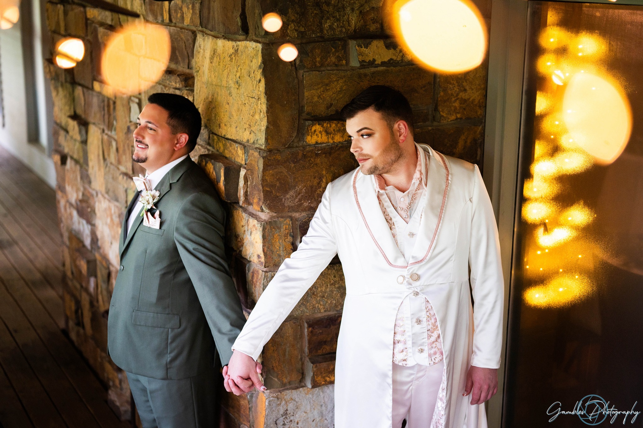 A couple holds hands on two sides of a stone wall, warm string lights frame the top of both people's heads as they look onward with anticipation. Photographed by Gambles Photography at a wedding at Affinity Riverside Estate in Nixa, Missouri