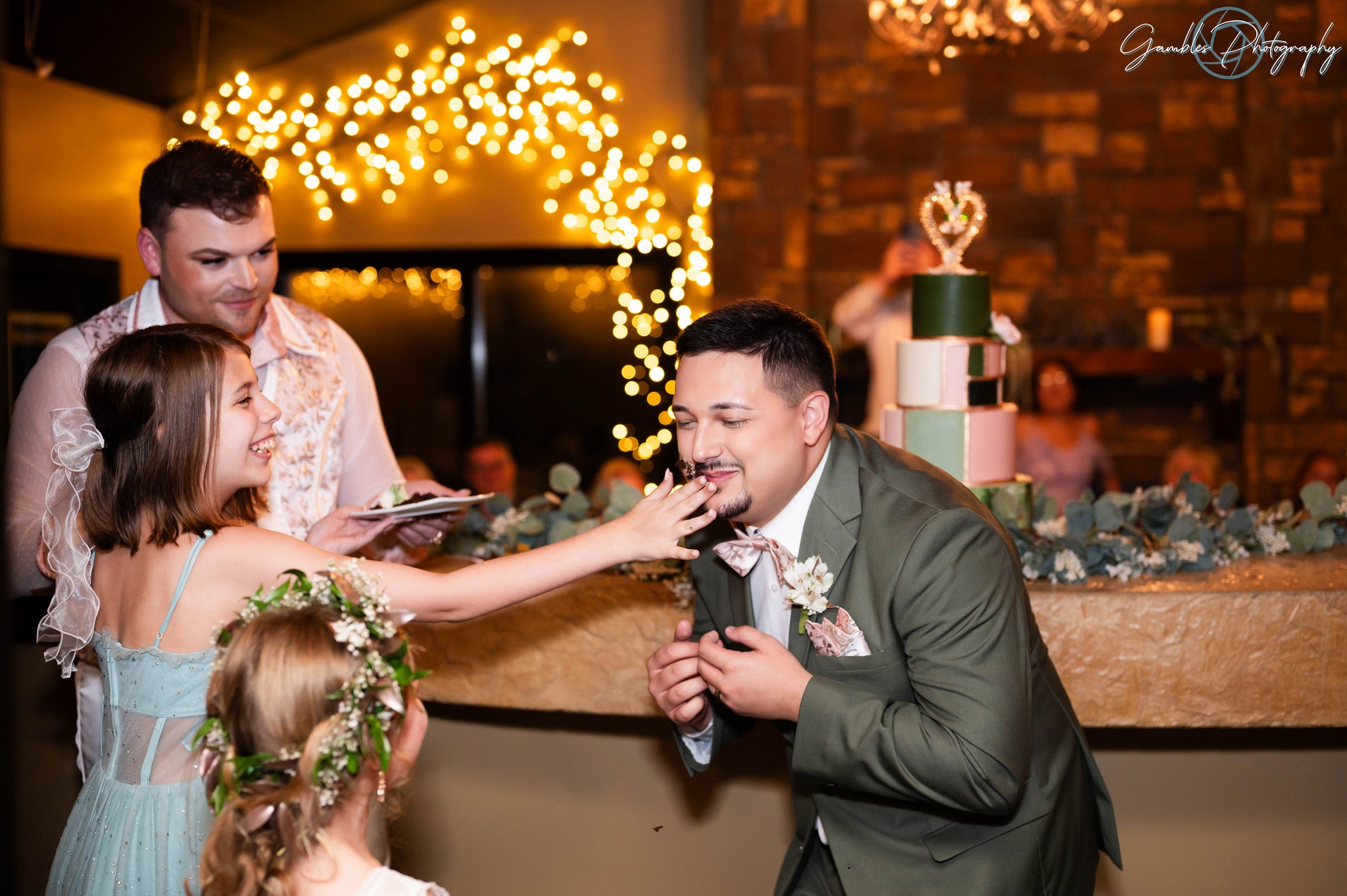 A flower girl touches a groom's face as he smiles during a wedding reception of Affinity Riverside Estate, photo by Gambles Photography