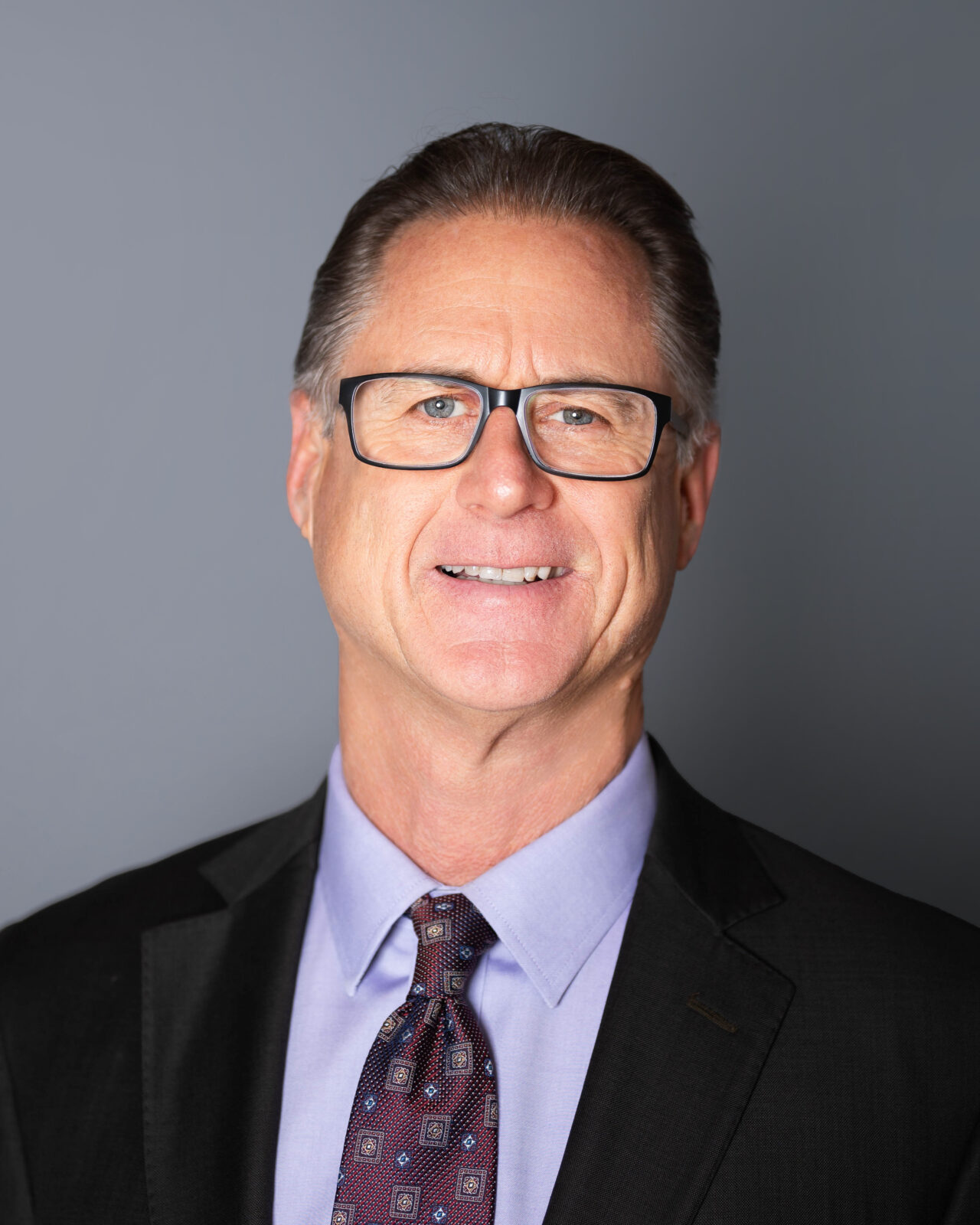 A man in glasses, a purple shirt, a dark purple tie, and black suit jacket smiles at the camera during headshots near Springfield, MO, by Gambles Photography.