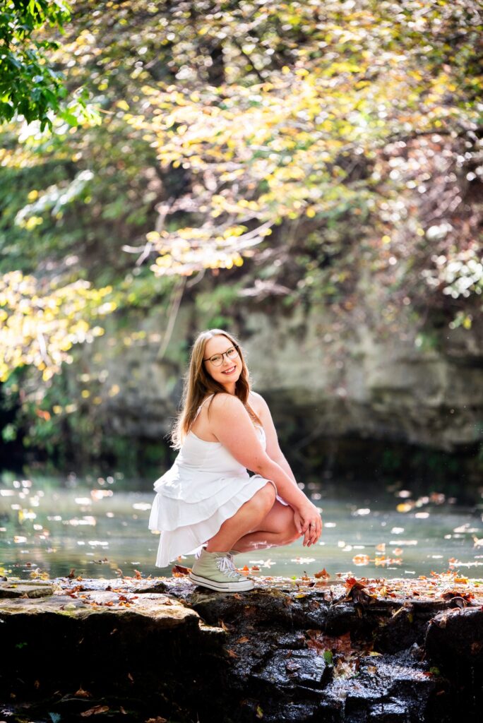 A woman in a white dress and sneakers crouches by the edge of a serene creek in Dogwood Canyon Nature Park, surrounded by autumn foliage. She smiles warmly at the camera, with natural light highlighting the peaceful woodland setting.