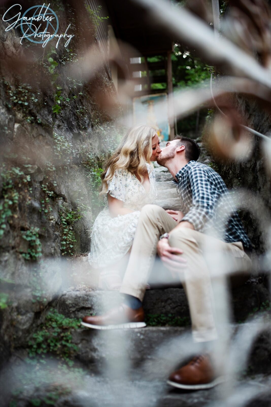 Sitting on dark grey stone steps and surrounded by ivy-colored stone walls, a couple kisses during a photoshoot in Eureka Springs, Arkansas. By Gambles Photography