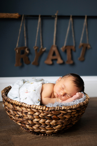 A baby sleeps in a wood basket with his name in wooden letters hung in the background. Taken during an infant photoshoot by Gambles Photography