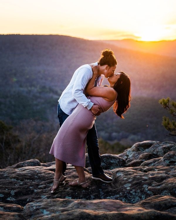 A lesbian couple kisses in Jasper, Arkansas, on top of a cliff with the red-orange sunset behind them. 