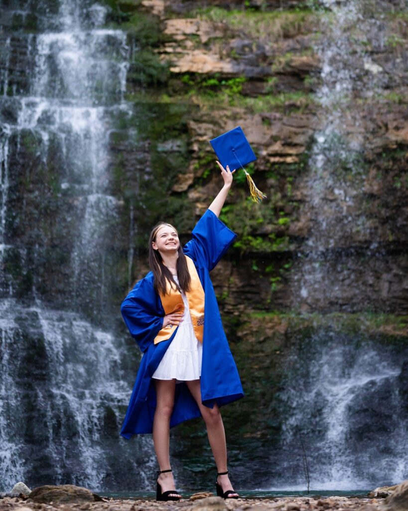 a high school graduate poses for senior photos in her stole, holding a blue grad cap above her head, in front of a waterfall in Jasper, Arkansas, photographed by Gambles Photography