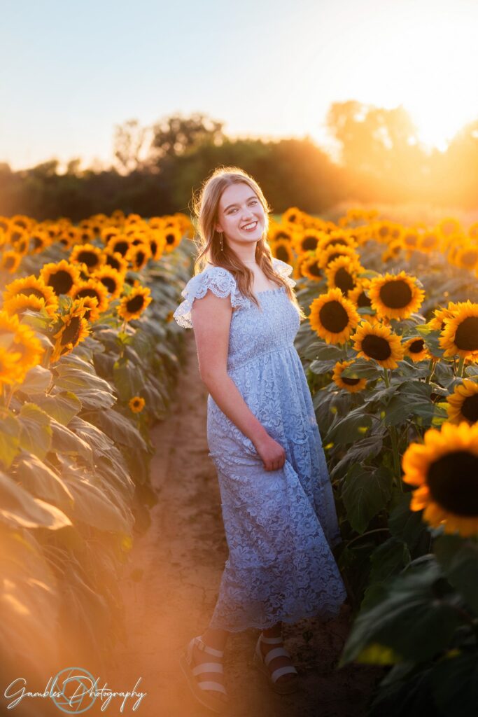 A young woman in a light blue lace dress smiles brightly as she stands in the middle of Pa's Posey's Garden at sunset. The golden light creates a warm, soft glow around her, complementing the vibrant yellow sunflowers in Cassville, MO.