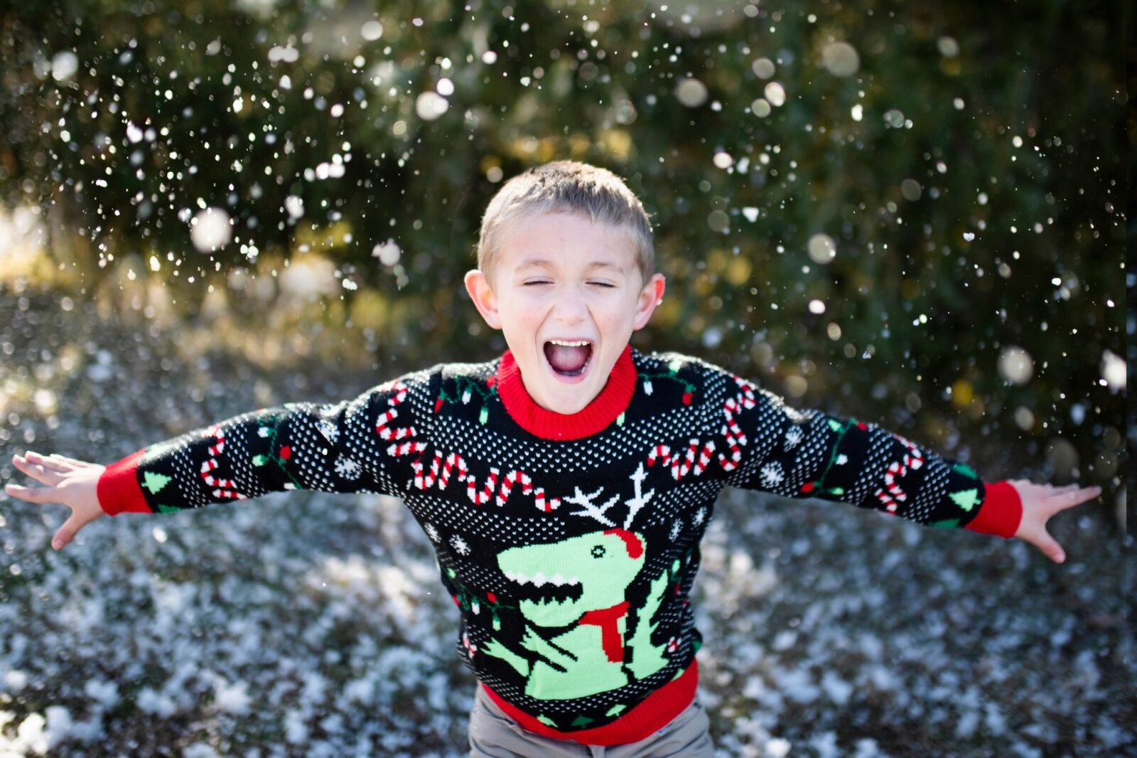 Child joyfully playing in light snow while wearing a dinosaur-themed Christmas sweater, arms outstretched.