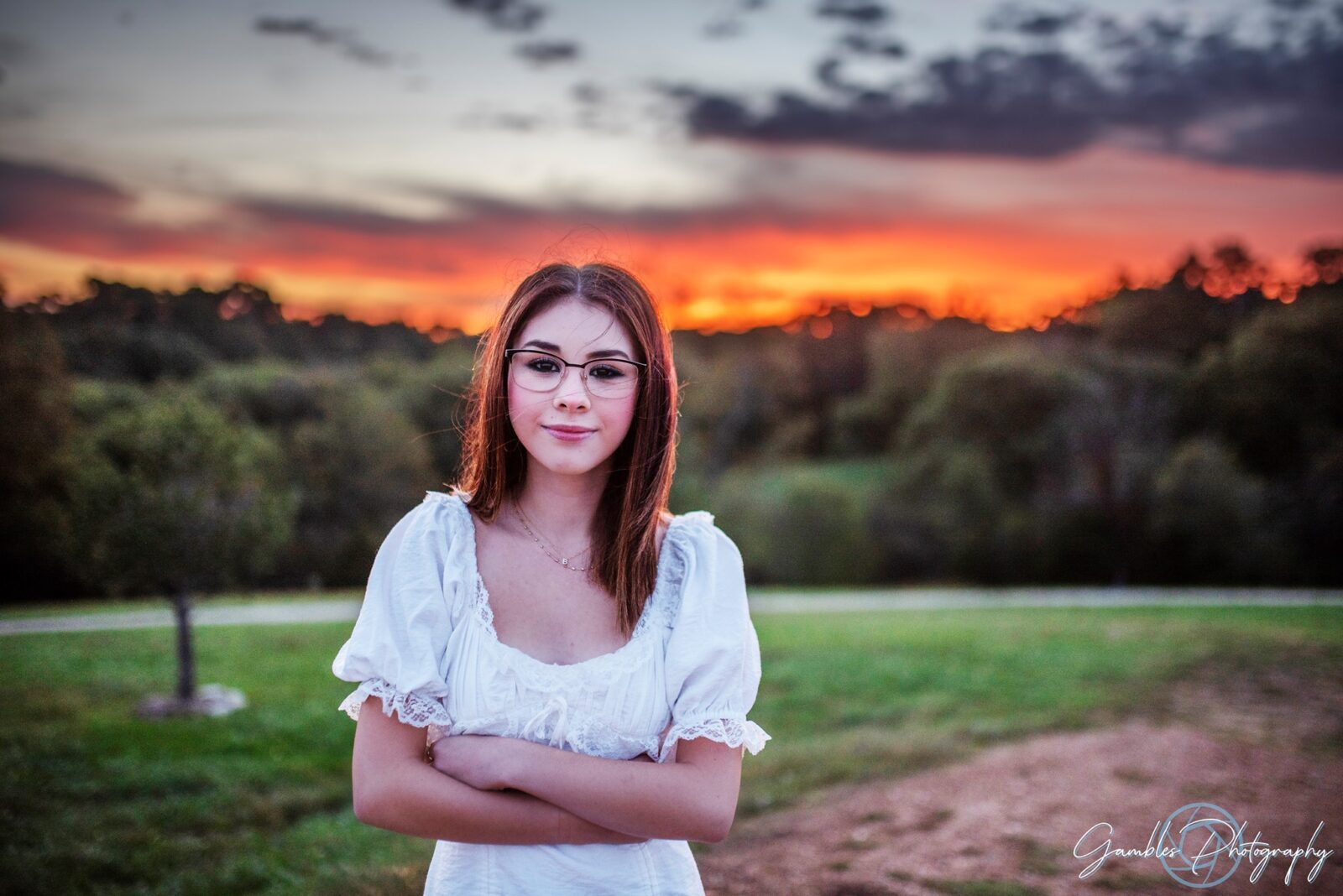 a young girl with glasses faces the camera, crossing her arms over her lacey, white dress as a red-orange sunrise bursts above the green trees and grass behind her. Photo by Gambles Photography