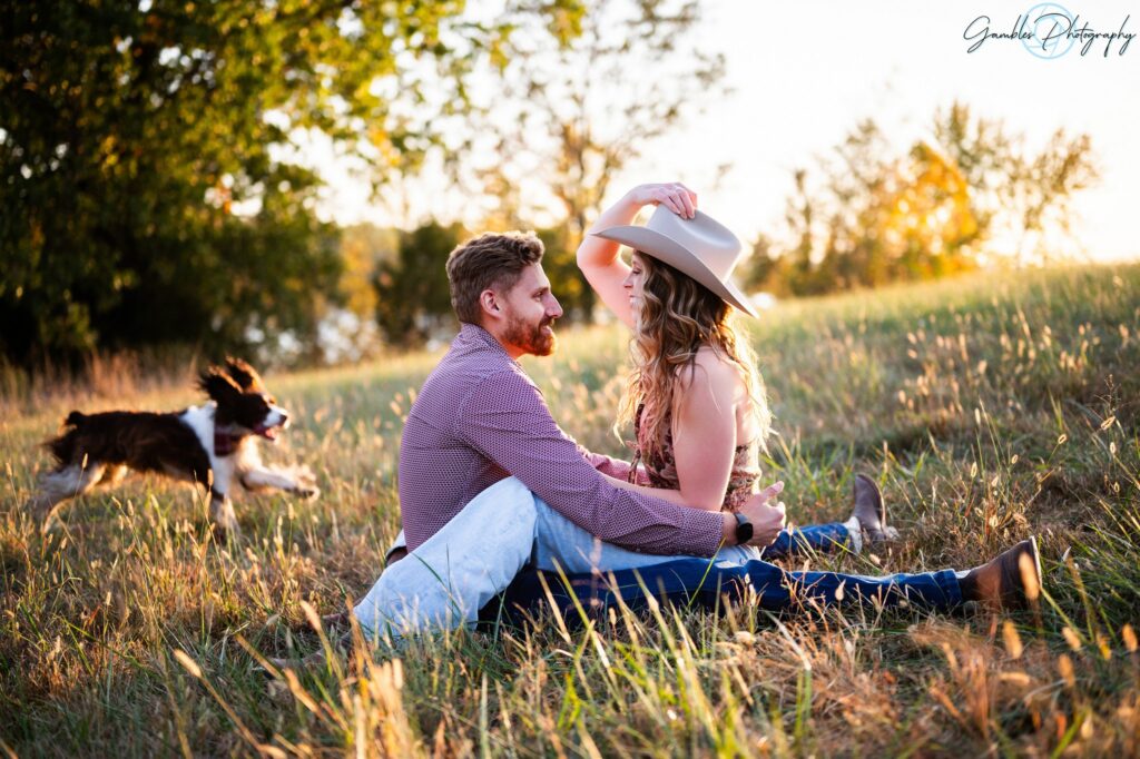 A couple sits facing each other, their legs wrapped around one another's waists, as the wife holds a white cowboy hat on to her head. The couple looks into each other's eyes and smiles.