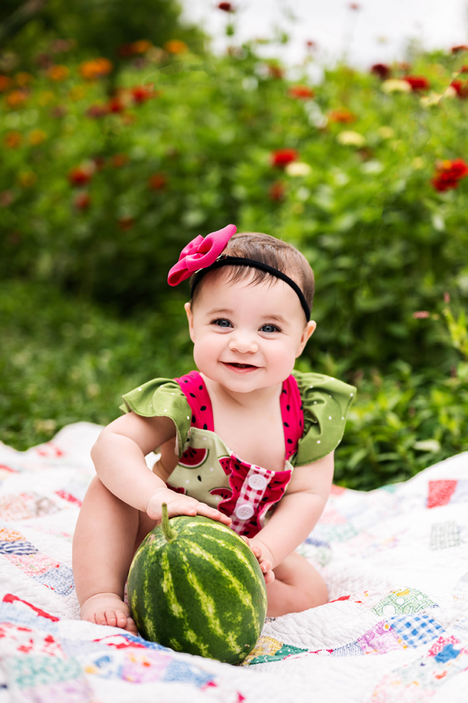 a toddler sits in a field of red flowers, dressed in an outfit to  match the watermelon she sits in front of.