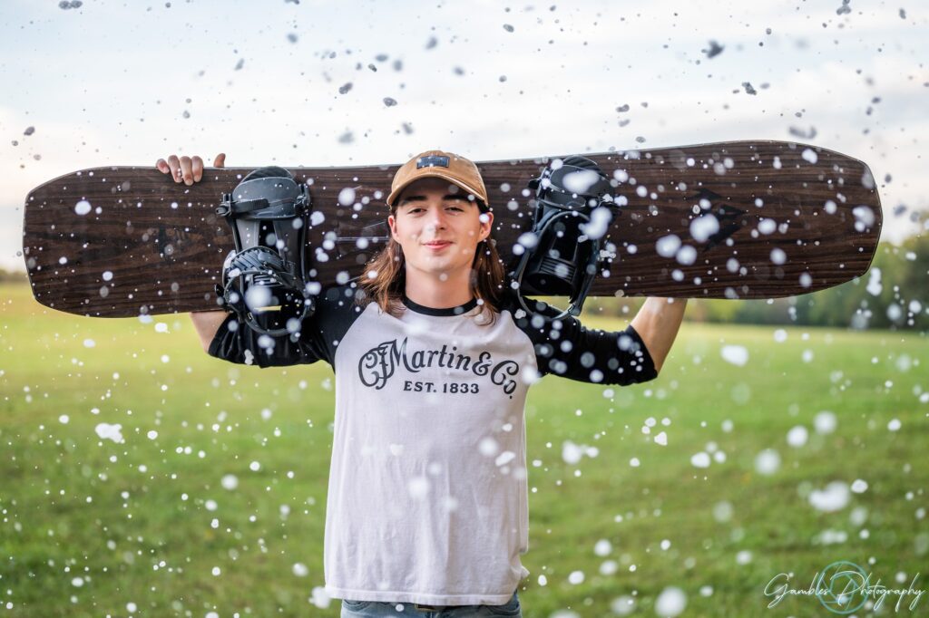 A high school senior poses with green grass in the background and fake snow in the foreground, smiling at the camera and using both arms to hold a snowboard behind his back. Taken during a senior photoshoot in Springfield, MO by Gambles Photography