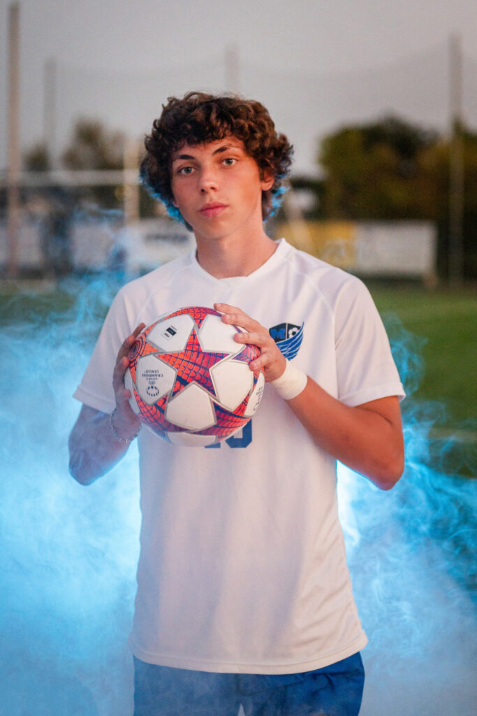 a high school senior holds a soccer ball with colored fog behind him during a Springfield, MO, senior portrait session by Gambles Photography