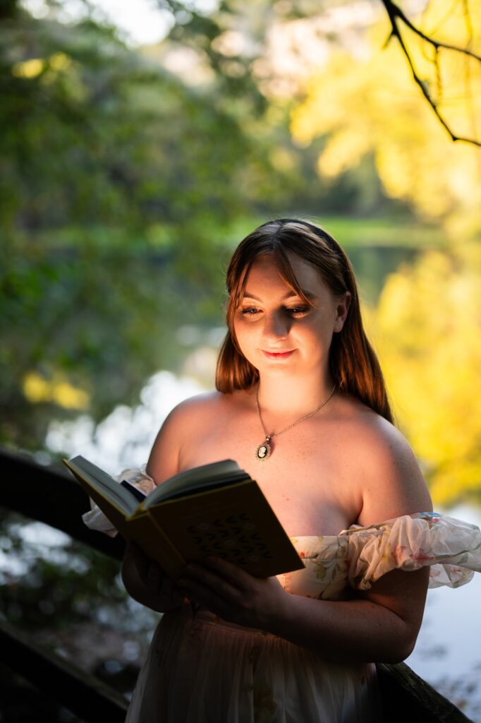 A photoshoot for Springfield, MO high school senior shows a woman smiling, reading a book that is illuminated with a lake behind her. Photo by Gambles Photography