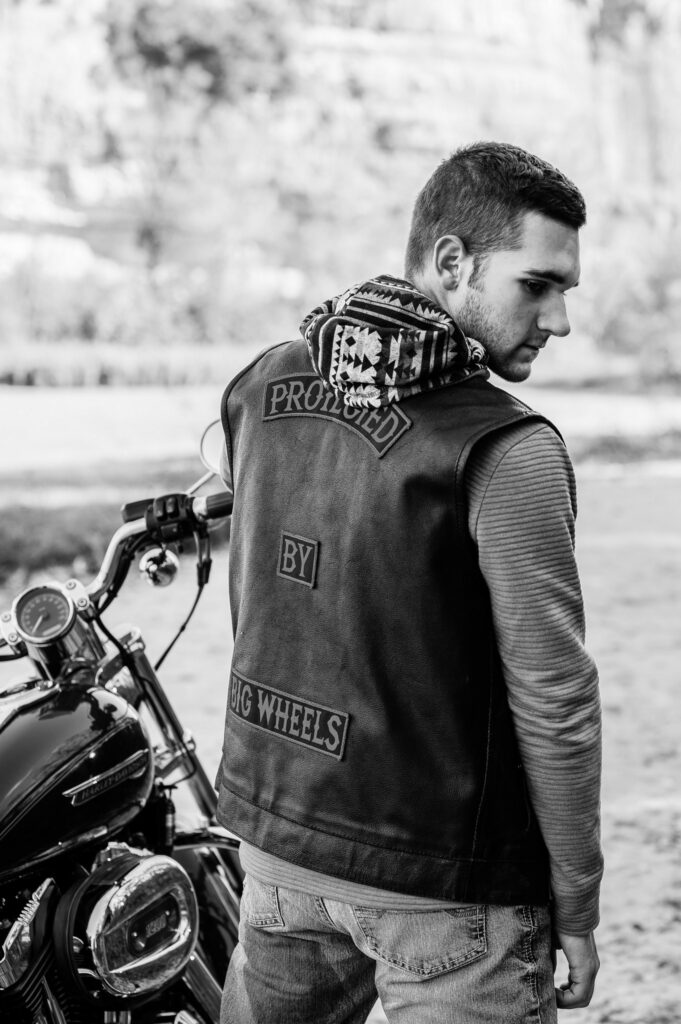 With a motorcycle in the background, a young man looks over his shoulder, wearing a leather vest that says 'Protected by big wheels' on the back. Photo by Gambles Photography